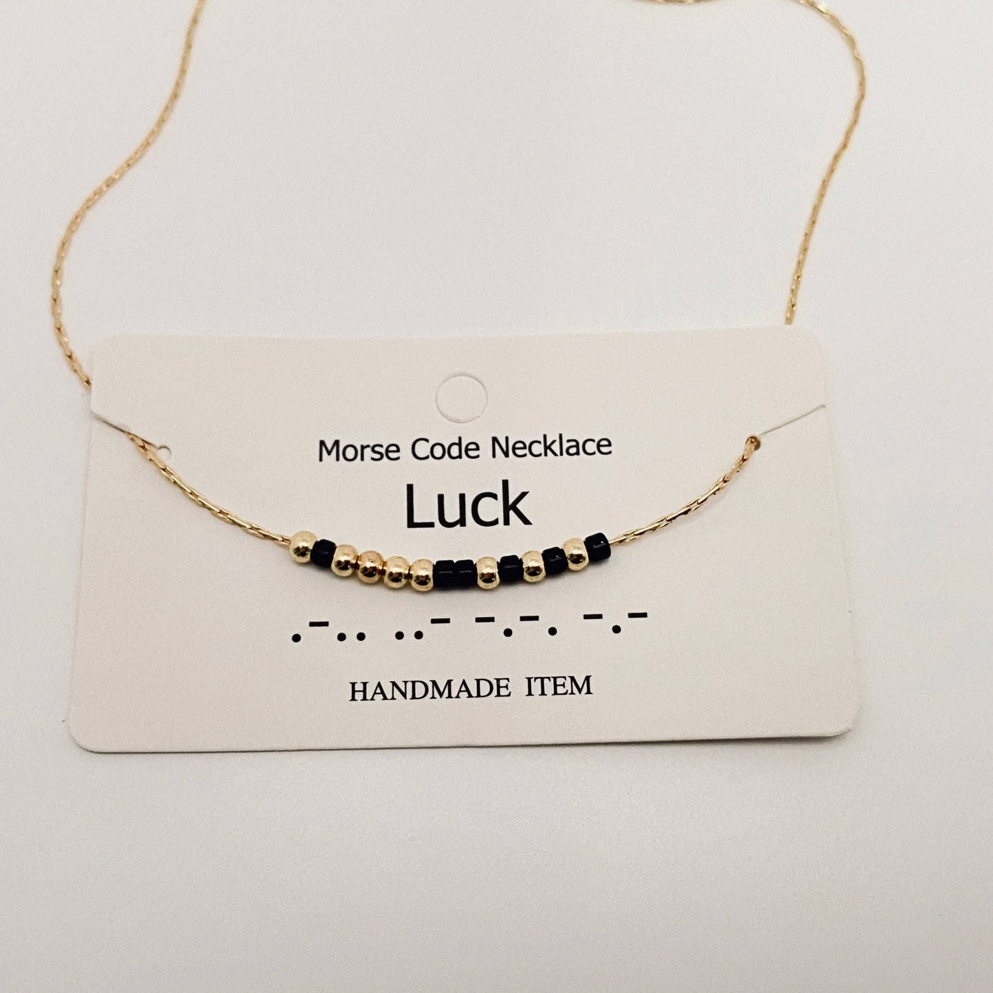 Morse Code Necklace labeled 'Luck' on a white card with gold chain and black beads.