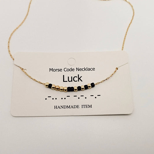 Morse Code Necklace labeled 'Luck' on a white card with gold chain and black beads.