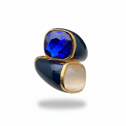 A close-up of a two-tone statement ring held on a person's fingers. The ring features a deep blue faceted gemstone on one side and a milky white cabochon gemstone on the other, both set in polished gold-tone metal with a glossy dark blue enamel inlay.