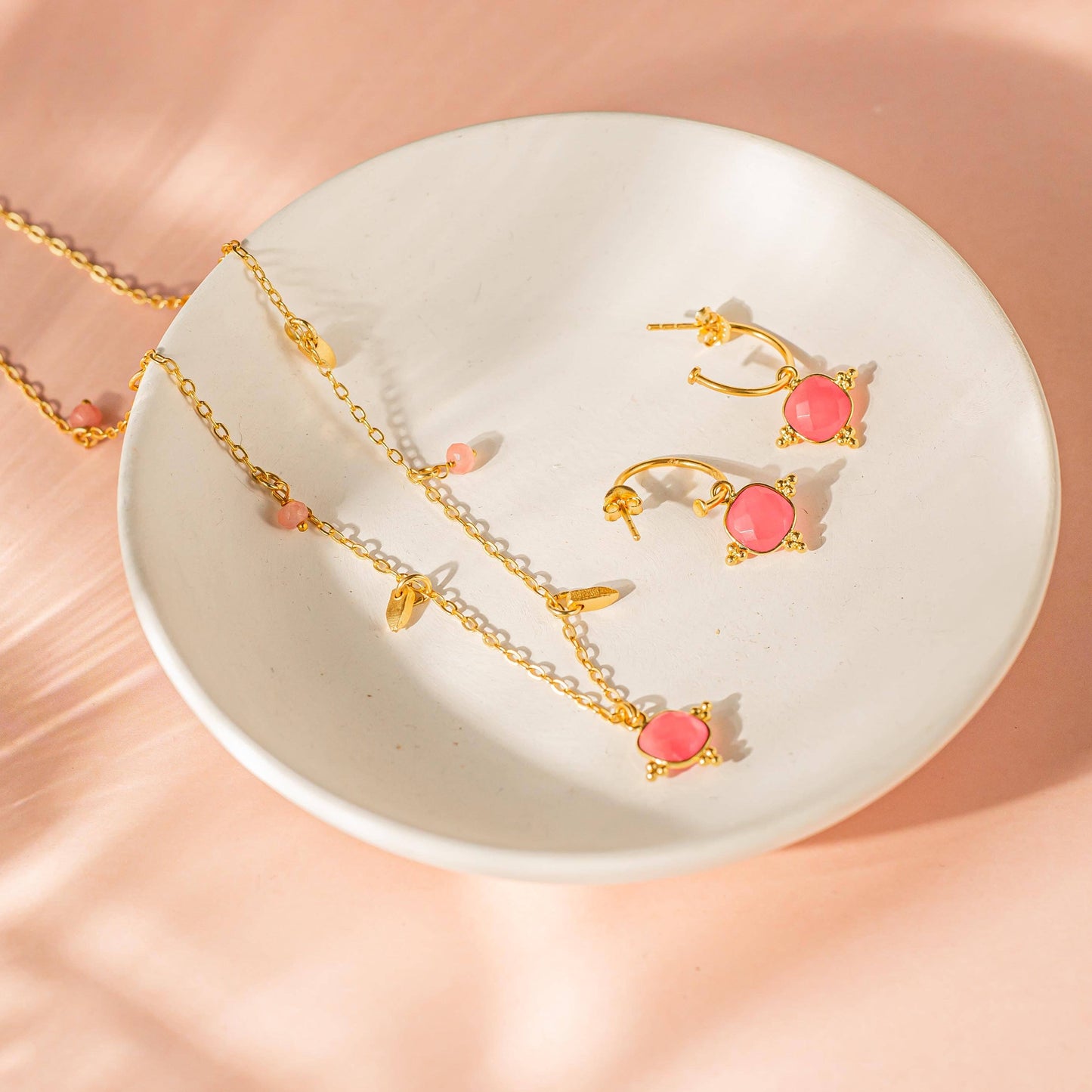 Gold necklace and earrings with pink stones on a white dish against a peach background