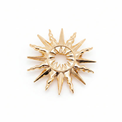 A gold-toned sunburst pendant or brooch with a circular center and radiating pointed rays, against a light neutral background.
