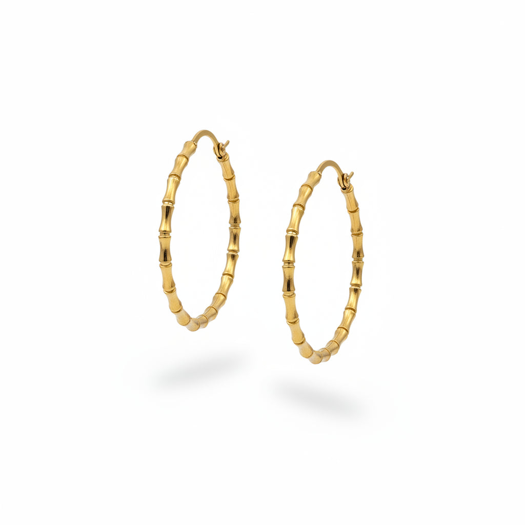 A pair of gold hoop earrings with a textured, beaded bamboo-like design, displayed on a person's hand over a gray textured background.