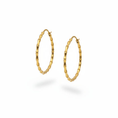 A pair of gold hoop earrings with a textured, beaded bamboo-like design, displayed on a person's hand over a gray textured background.